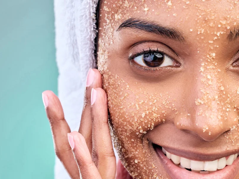 A woman with a towel on her head applies brown exfoliating scrub to her face, showcasing a skincare routine.