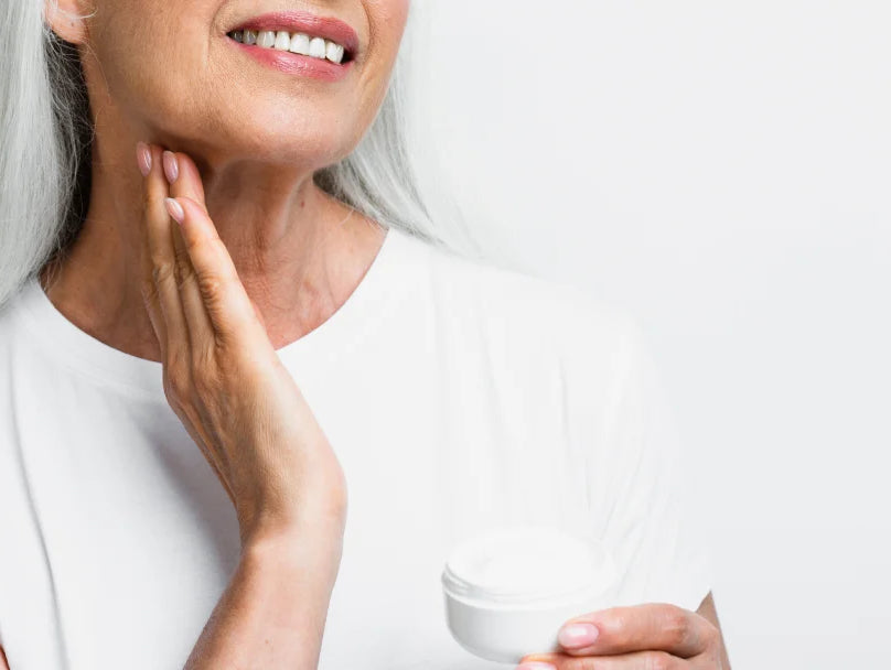 A woman with gray hair holds a jar of anti-wrinkle cream, smiling as she promotes skincare for mature skin.