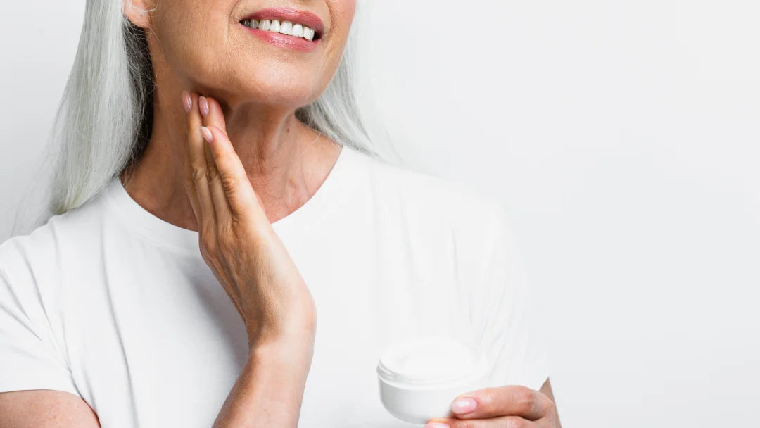 A woman with gray hair holds a jar of anti-wrinkle cream, smiling as she promotes skincare for mature skin.